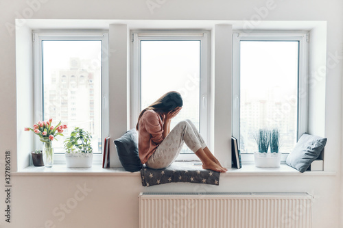 Cuadros en Lienzo Sad young woman crying about something while sitting on the window sill at home