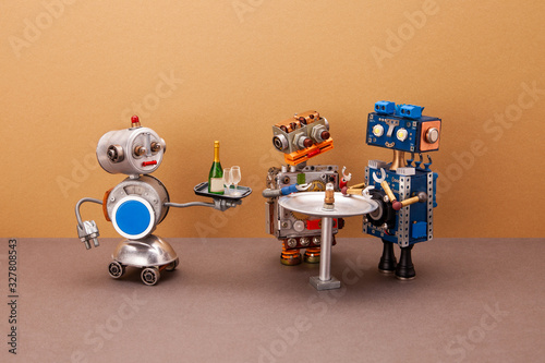 A four-wheeled robot waiter carries a tray with glasses and a bottle of champagne for two guests of robots who are waiting for their order at the restaurant table. Brown wall background.