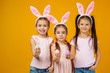© producer - happy cute little child girls with pink bunny ears holding painted Easter eggs on studio yellow background. Easter day