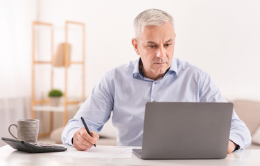 Serious senior man using laptop and writing