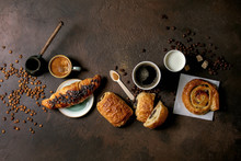 Variety Of Traditional French Puff Pastry Raisin And Chocolate Buns, Croissant With Various Cups Of Coffee And Milk, Cezve, Recycled Wooden Spoon Of Sugar Over Dark Texture Background. Flat Lay, Space