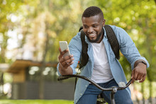 Happy Black Man Riding Bicycle Outdoors And Using Smartphone