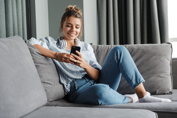 Wall Mural - Image of smiling nice woman using cellphone while sitting on sofa