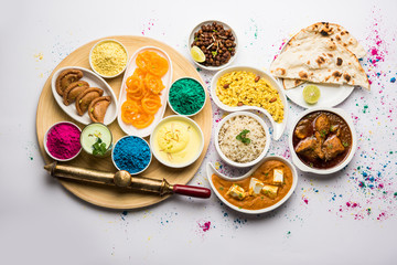 Happy Holy concept  showing Indian assorted lunch food like chicken, paneer butter masala, naan, jeera rice, black chana fry, jalebi, fujiyama, thandai and Farsan with holi colours and pichkari 