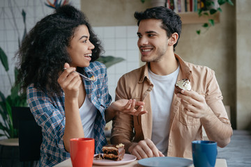 Happy multiracial friends communication together. Young lovely couple laughing, sitting in cafe. Beautiful African American woman and Indian man drinking coffee, eating tasty cake. First date concept