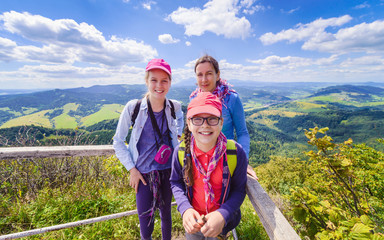 Happy family - mother and two daughters - on a hiking trip to the mountains - on top of a mountain with blue sky and clouds in the background