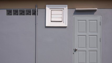 Wall Mural - Front view of gray wooden door, square ventilation shutter, glass blocks and fluorescent light bulb with PVC pipe on gray cement wall with brown stripe, image for home exterior decoration concept