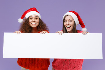 Wall Mural - Funny european african american women friends in Christmas hat isolated on violet purple background. Happy New Year celebration holiday. Mock up copy space. Hold blank sign board with place for text.