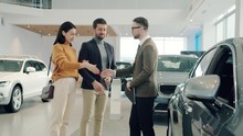 Slow Motion Of Young Couple Man And Woman Buying Car Shaking Hands With Agent Hugging Taking Keys Enjoying Successful Deal. People And Shopping Concept.