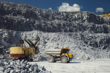 Wall Mural - Mining dump truck drives off from the excavator after loading in a quarry for the extraction of limestone on the background of rocky terrain and blue sky in sunny weather.