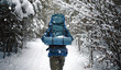 Back view of outdoorsman wearing big snow-clad backpack walking on winter forest path
