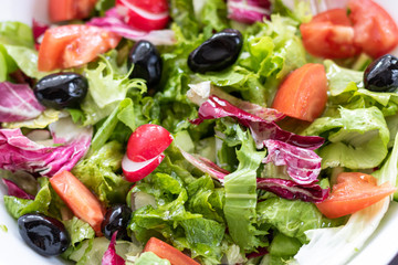 Green vegan salad from green leaves mix and vegetables in white bowl.Summer Easter diet salad dish of lettuce, radicchio, Lollo Rossa, endives, radishes, green onions, cucumbers, olives and tomatoes