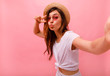 © ClaudiK - Portrait of a happy cheerful girl in summer hat showing peace gesture and making cute selfie isolated over pink background. Copy space