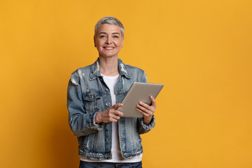 Smiling mature woman with digital tablet in hands on yellow background