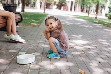 cute little girl sitting and drawing with chalk on asphalt in park.