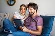 © PhotoAlto - Smiling young couple using digital tablet while relaxing on sofa at home
