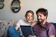 © PhotoAlto - Smiling young couple using digital tablet while relaxing on sofa at home