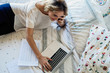 © PhotoAlto - Young woman talking on smartphone while using laptop on bed in bedroom