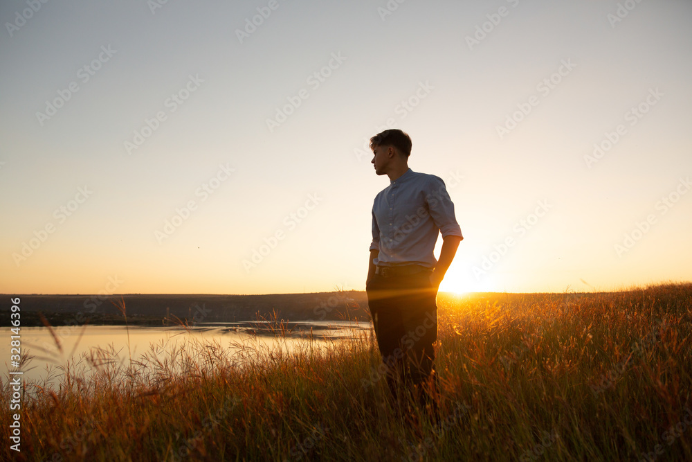 Man standing and enjoying the beautiful sunset over a wide river valley. Young man in  blue shirt and black pants standing and looking to river. - obrazy, fototapety, plakaty 