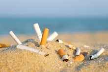Cigarette Butts In Yellow Sand On A Sea Beach On The Coast Against A Background Of Blue Sky And Sea. The Problem Of Humanity.  Cigarette Smoking, A Bad Habit. Nicotine Addiction. Garbage