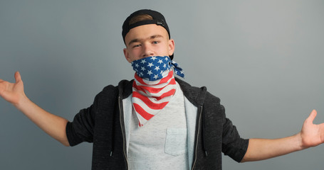 Wall Mural - Portrait of young protester teenager boy hides his face behind flag USA aggressively shrugs his hands at opponent on grey background. Aggression. Protests. Independence. Freedom, rebel