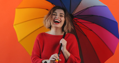 Wall Mural - Rainbow umbrella. Portrait of happy smiling young sexy girl rotating rainbow umbrella in red sweater on red background slow motion. Pleasure. Emotions of people. Monotone. Copy space