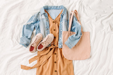 Wall Mural - Blue jean jacket and beige dress, sneakers with tote bag on white bed. Women's stylish autumn outfit. Trendy clothes. Flat lay, top view.
