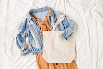 Wall Mural - Blue jean jacket and beige dress with tote bag on white bed. Women's stylish autumn outfit. Trendy clothes with white eco bag mockup. Flat lay, top view.