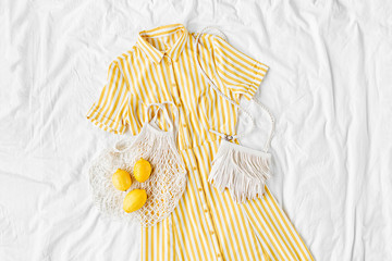 Yellow dress with stripes with eco bag and lemons on white bed. Women's stylish  summer outfit. Trendy clothes. Flat lay, top view.