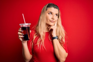 Poster - Young beautiful blonde woman drinking cola fizzy beverage using straw over red background serious face thinking about question, very confused idea