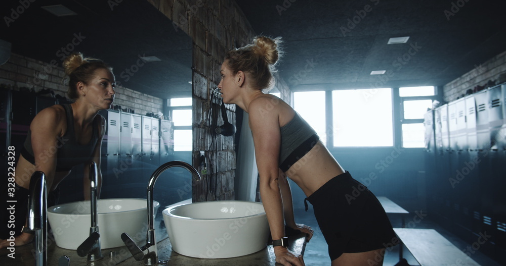 Attractive tall fit blonde woman taking shirt off, looking in the mirror in dark gym locker room exhausted slow motion. - obrazy, fototapety, plakaty 