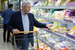 Mature man choosing dairy products in store. Serious man with shopping trolley looking at shelves in refrigerator while buying goods in supermarket. Shopping concept