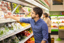 Young Man Shopping In Grocery Store. Side View Of Focused Man And Woman Holding Shopping Baskets And Choosing Fresh Fruits And Vegetables In Supermarket. Shopping Concept