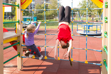 Two Happy Caucasian Girls Having Fun On Playground In Summer, Climbing The Rope Net. The Older Girl Is Hanging Upside Down
