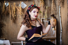 Portrait Of A Young, Attractive Witch Sitting At A Table With Magic Books And Scrolls Against A Background Of Alchemical Ingredients, Flasks, Bottles, And Dried Herbs.