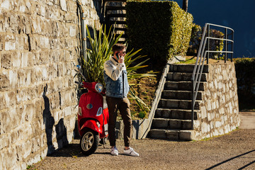 Handsome young man with mobile phone standing near red motorcycle in the city and speak. Nice attractive stylish trendy confident man holding in hands phone.