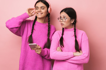 Wall Mural - Image of upset girl standing by her happy female friend with cellphone