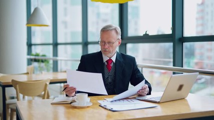 Wall Mural - Businessman is reading a printed report or document while sitting at the table, working on paper work with laptop at the table. Man looking at documents. Business video.