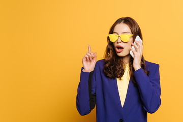 amazed, stylish girl in yellow glasses showing idea sign while talking on smartphone isolated on yellow