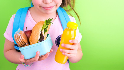 Young schoolgirl with lunch box on a green background. Little girl with a school backpack and a set of food for a snack. Free space for text.