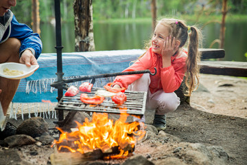 Man and his little daughter having barbecue in forest on rocky shore of lake, making a fire, grilling bread, vegetables and marshmallow. Family exploring Finland. Scandinavian summer landscape. 