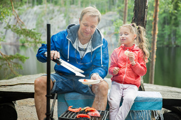 Man and his little daughter having barbecue in forest on rocky shore of lake, making a fire, grilling bread, vegetables and marshmallow. Family exploring Finland. Scandinavian summer landscape. 