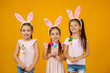 © producer - happy cute little child girls with pink bunny ears holding painted Easter eggs on studio yellow background. Easter day