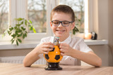 Fototapeta  - Chłopiec w wieku szkolnym siedzi przy stole i szeroko się uśmiecha. Wkłada do skarbonki w kształcie piłki nożnej banknot o nominale 20 euro.