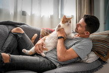 Hombre Joven Recostado En Un Sofa, Toma Entre Sus Brazos A Un Gato Blanco Y Marron Y Lo Mira Con Cariño