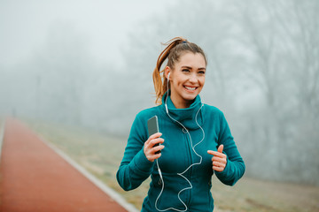 Wall Mural - Young fit girl is listening to music from her smartphone while running outside on foggy day.