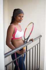 Wall Mural - Portrait of an attractive young brunette dark skinned Indian Bengali girl in western sportswear and a tennis bat in a balcony in white background. Indian lifestyle