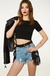 © RedUmbrella&Donkey - A brown-haired lady in a black top and denim shorts is wearing a black leather belt with straps, fringe, steel rivets, rings and a buckle. The lady with a biker jacket is posing on the gray backdrop.