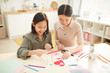 © AnnaStills - Young Asian woman and her teen sister creating handmade Mothers Day holiday card with hearts, horizontal high angle shot, copy space