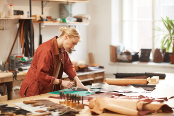 Wall Mural - Horizontal side view portrait of modern female artisan doing leather craftwork standing at table in her workshop